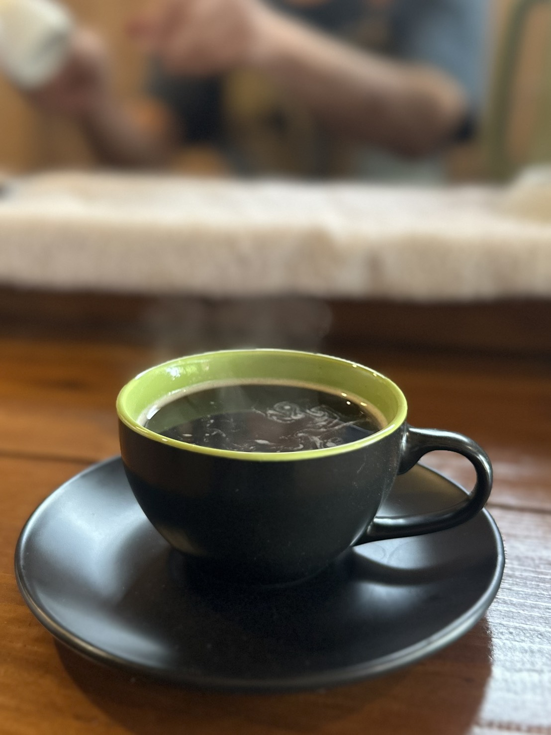 木のカウンターに置かれた黒いカップとソーサー。緑色の縁が印象的なカップに温かいコーヒーが注がれ、湯気が立っている。